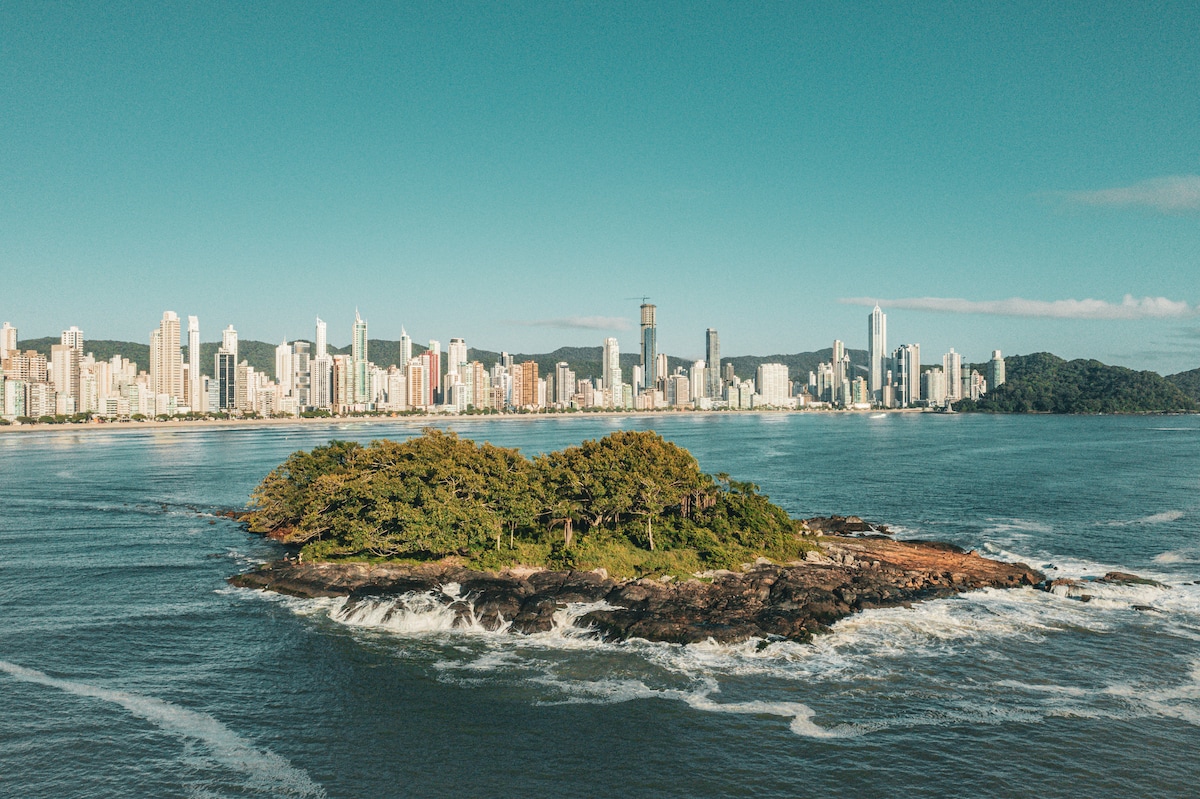 An aerial view captures a small island surrounded by calm waters, featuring lush greenery and rocky shorelines. The city skyline of Balneário Camboriú rises in the background, showcasing a mix of modern skyscrapers and natural beauty under a clear blue sky.