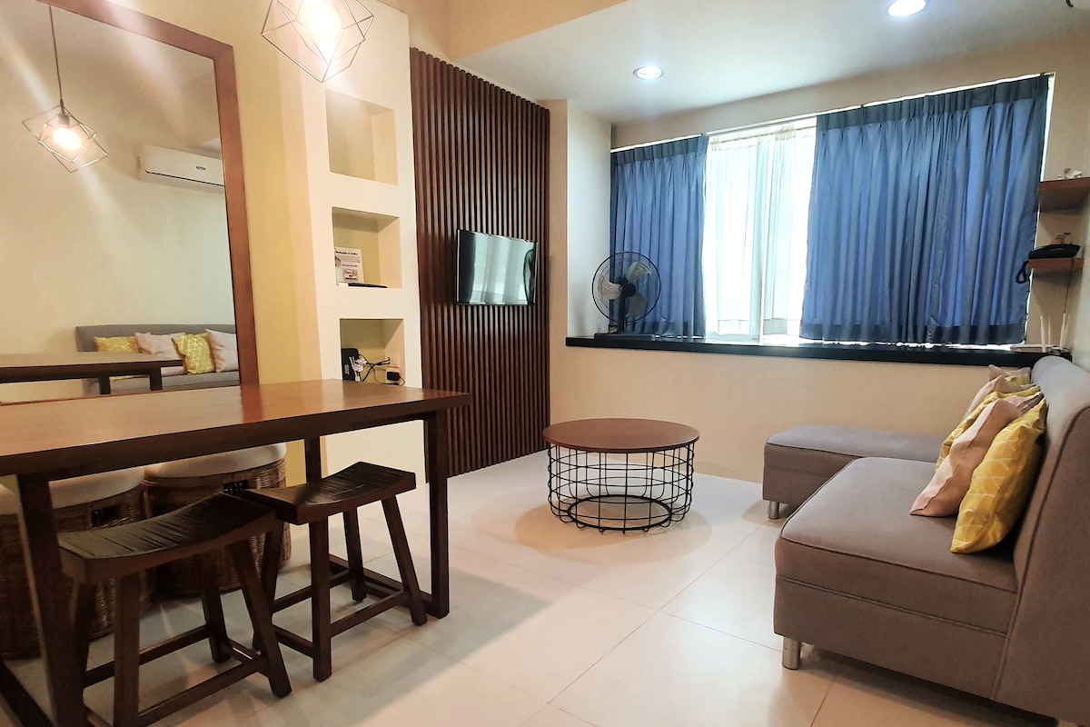 A bright living area features a comfortable gray sofa and a round coffee table with a wire design. A dining table with stools is positioned beside a wall-mounted television. Natural light enters through the large window, adorned with blue curtains.