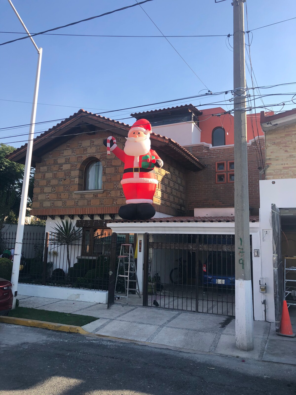 An inflatable Christmas decoration of Santa Claus is prominently displayed on the rooftop of a residential house. The home features textured stonework and a tiled roof, with greenery visible in the front yard. Utility lines run along the side of the structure.