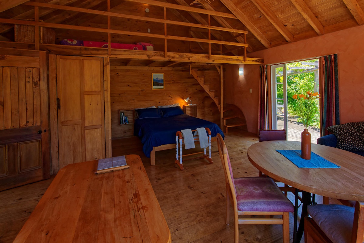 An open-plan living space features wooden beams and natural light. A spacious bed is positioned against one wall, with a cozy seating area nearby. A dining table is set with a blue table runner, and large doors lead to a private deck overlooking the orchards.