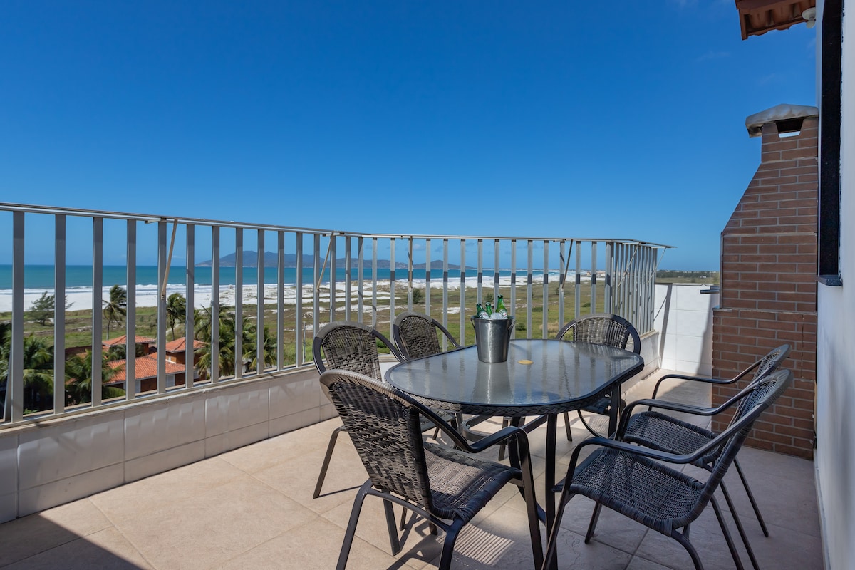 A spacious terrace is presented with a glass-topped dining table surrounded by six sturdy chairs. A charcoal grill is positioned nearby, with views of the ocean and coastline in the background, complemented by clear blue skies.