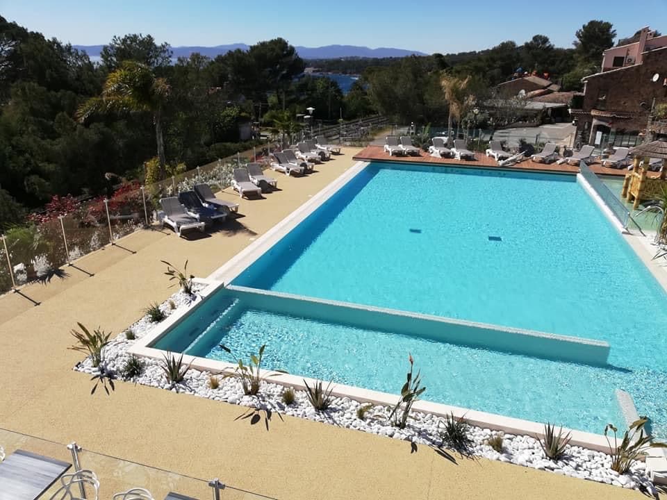 A spacious outdoor swimming pool is surrounded by landscaped areas featuring white stones and various plants. Sun loungers are positioned along the poolside, with a scenic view of distant hills and the sea.