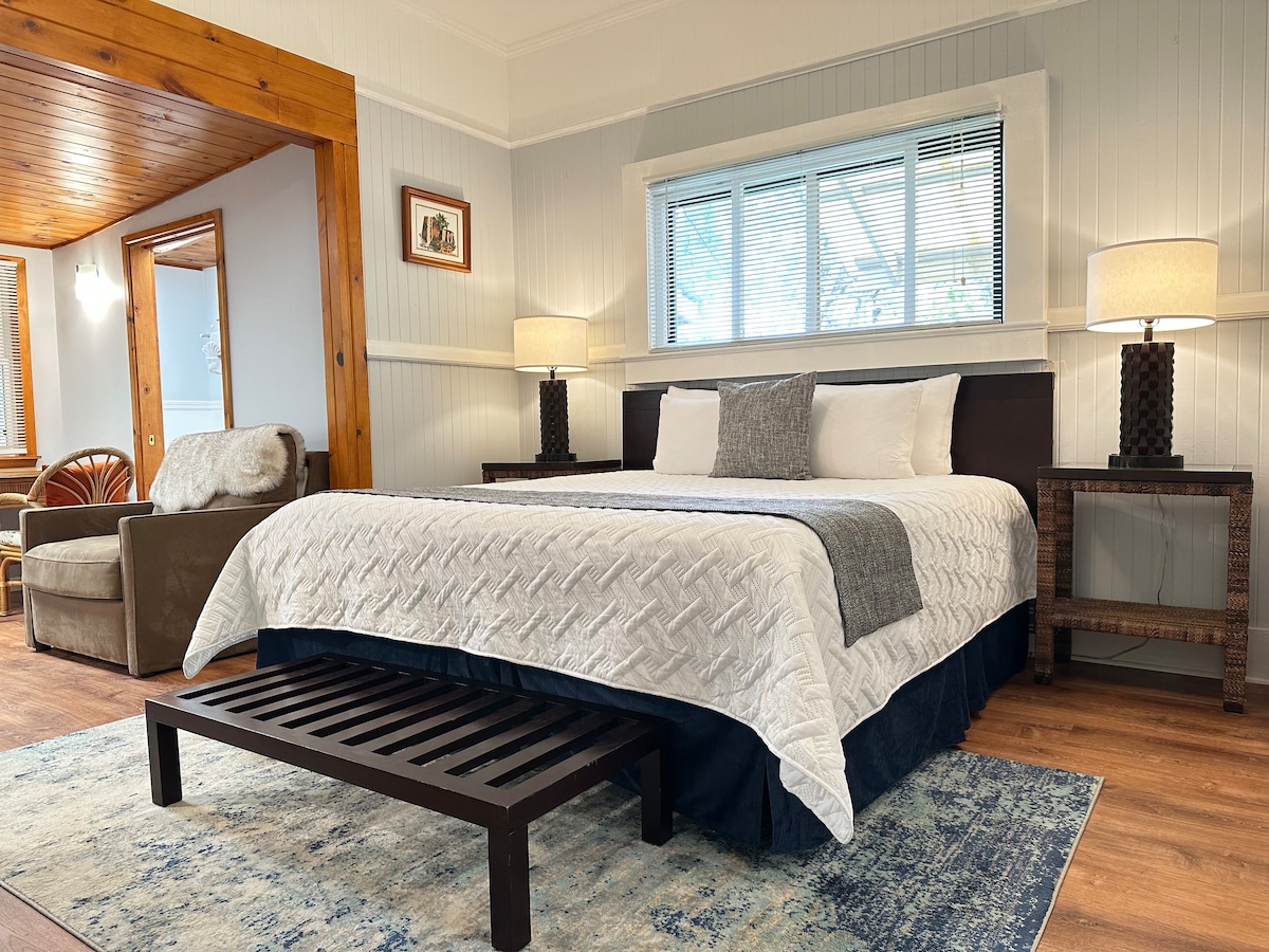 A spacious bedroom features a king bed with a textured white quilt and decorative pillows. A cozy pull-out sofa chair is positioned nearby. Light from a window illuminates the room, complemented by two bedside lamps. A wooden bench rests at the foot of the bed.
