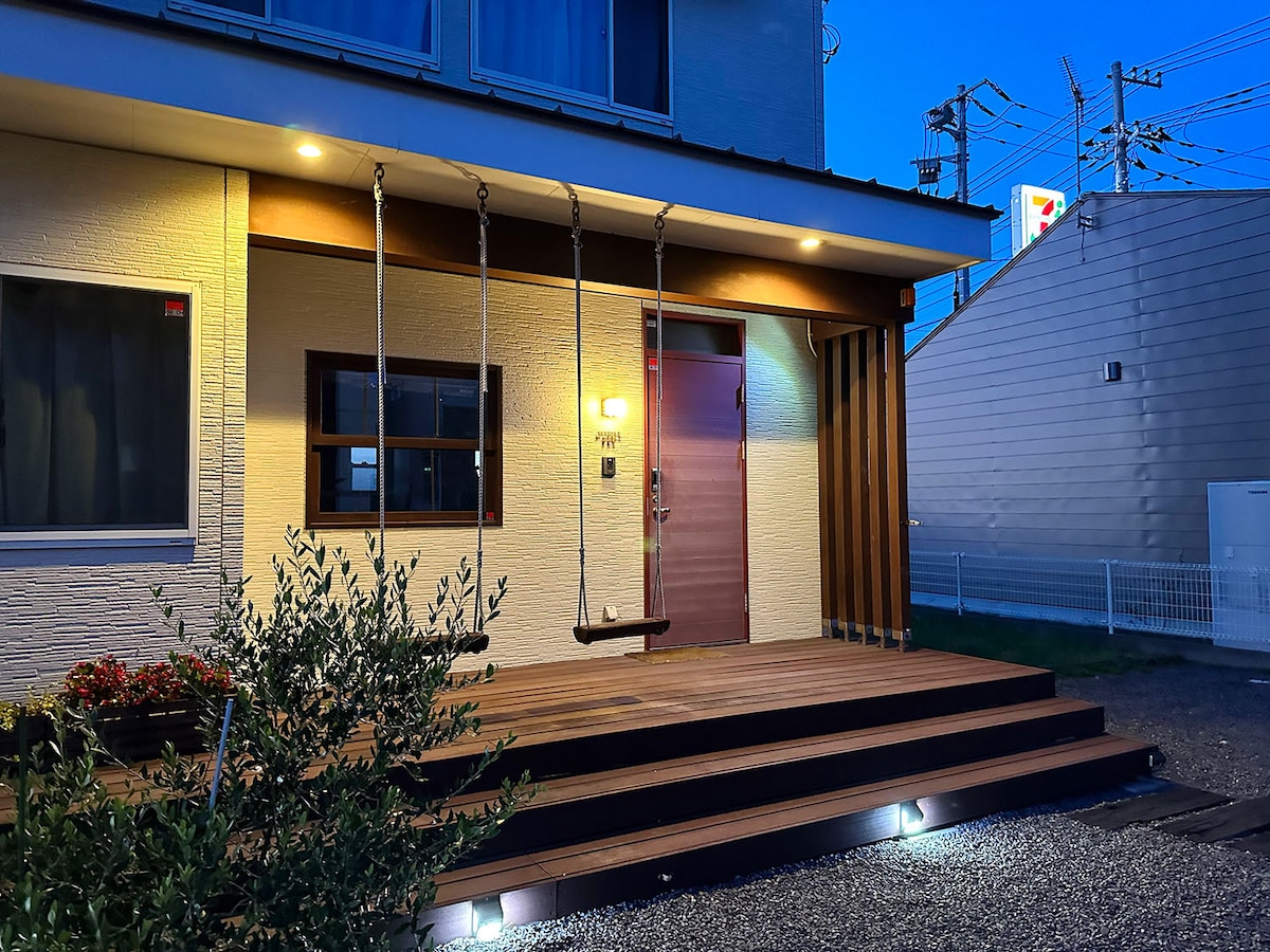 The exterior of the home features a welcoming entrance with a wooden porch and three steps leading to the door. A swing hangs from the porch, adding a playful element. Soft lighting highlights the porch area, while nearby greenery frames the view.
