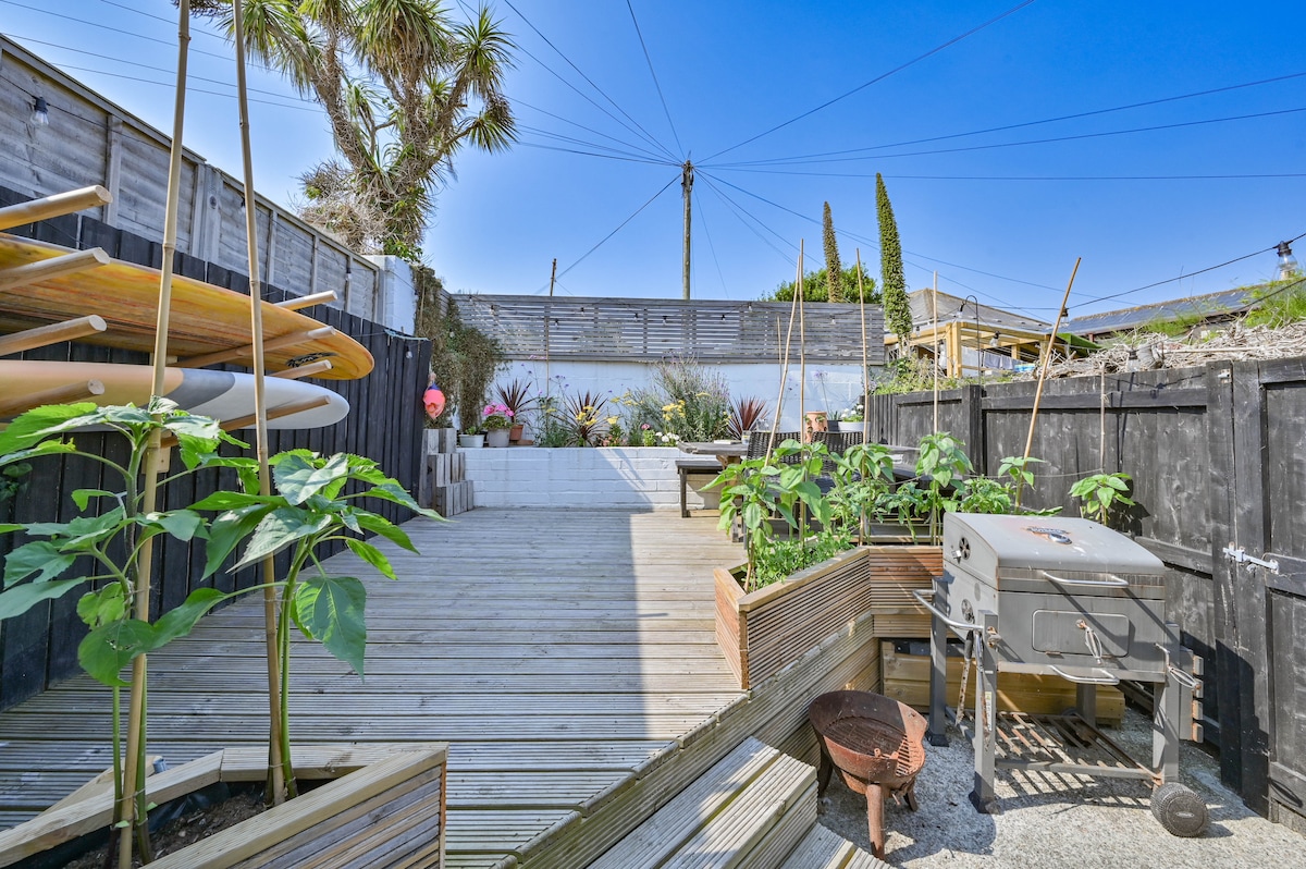 An outdoor space features a wooden deck surrounded by vibrant plants and surfboards. A barbecue sits adjacent to a fire pit, and built-in plant boxes are visible, creating an inviting area for relaxation and outdoor dining.