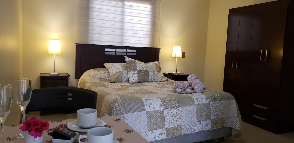 A spacious bedroom is visible featuring a large bed covered with a patterned quilt. Soft lighting is provided by two bedside lamps. A dark wooden wardrobe and a small coffee table with cups are also seen. Natural light enters through a window with light blinds.
