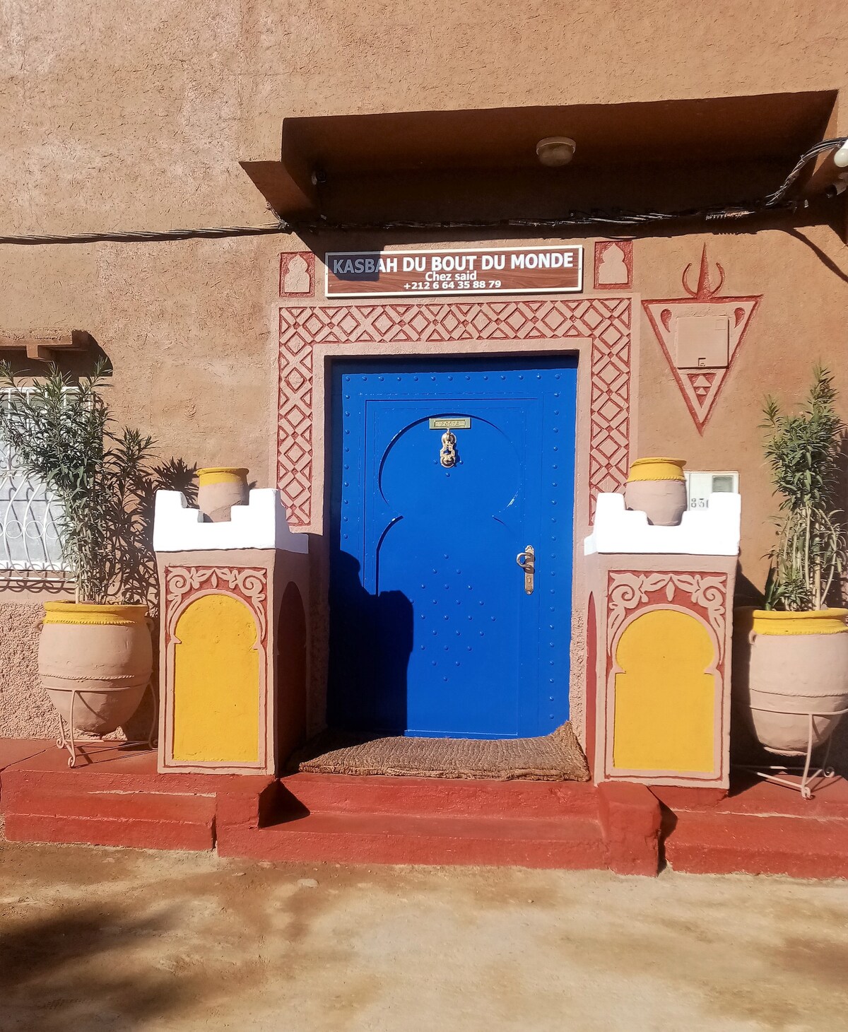 A vibrant blue door framed by decorative stonework leads to the kasbah, with accent features including terracotta planters. A rugged, earth-toned façade complements the entrance, which is adorned with tribal patterns. A welcome sign is positioned above the door, indicating the name of the kasbah.