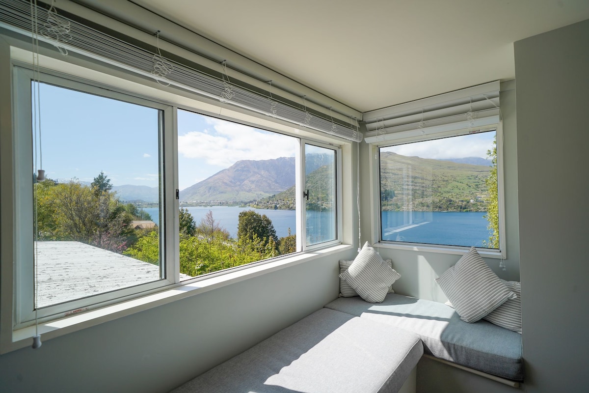 A bright nook features large windows offering views of the lake and mountains. Soft cushions are placed on a built-in seating area, inviting relaxation while enjoying the scenery. Natural light floods the space, enhancing the fresh and serene ambiance.