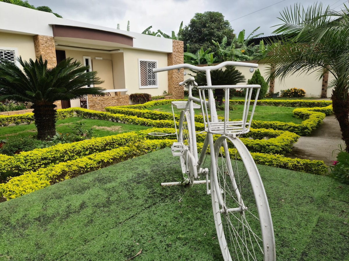 A white bicycle with a basket is positioned on green grass, serving as a decorative element. Lush flowerbeds with vibrant yellow and green plants surround the entrance to the house, offering a welcoming view that complements the tropical atmosphere.