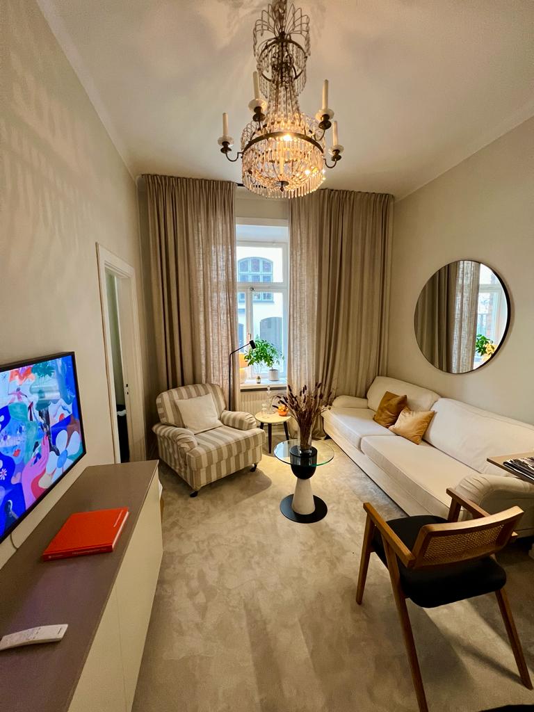 The lounge area features a white sofa and a light-colored armchair with stripes, complemented by a small table. A large mirror reflects natural light from the window, while a TV is mounted on a light-toned cabinet. Soft curtains frame the window, adding warmth to the space.