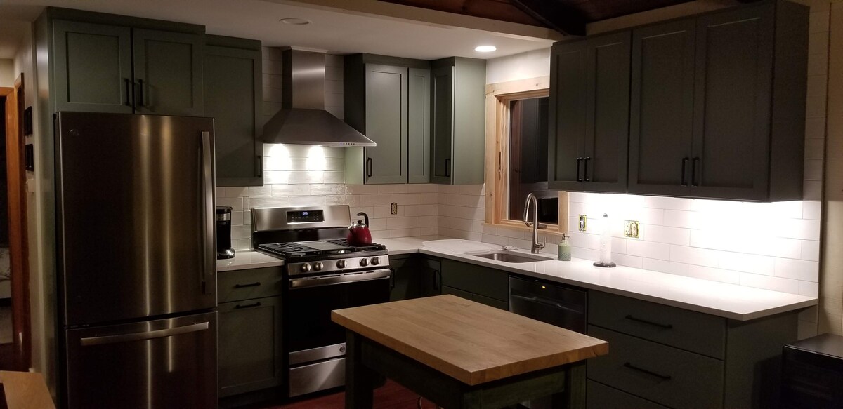 A modern kitchen is equipped with dark green cabinetry and stainless steel appliances. A large butcher block island provides additional workspace. Soft lighting illuminates the countertops, while a window allows for natural light. The design combines functionality with a clean aesthetic.
