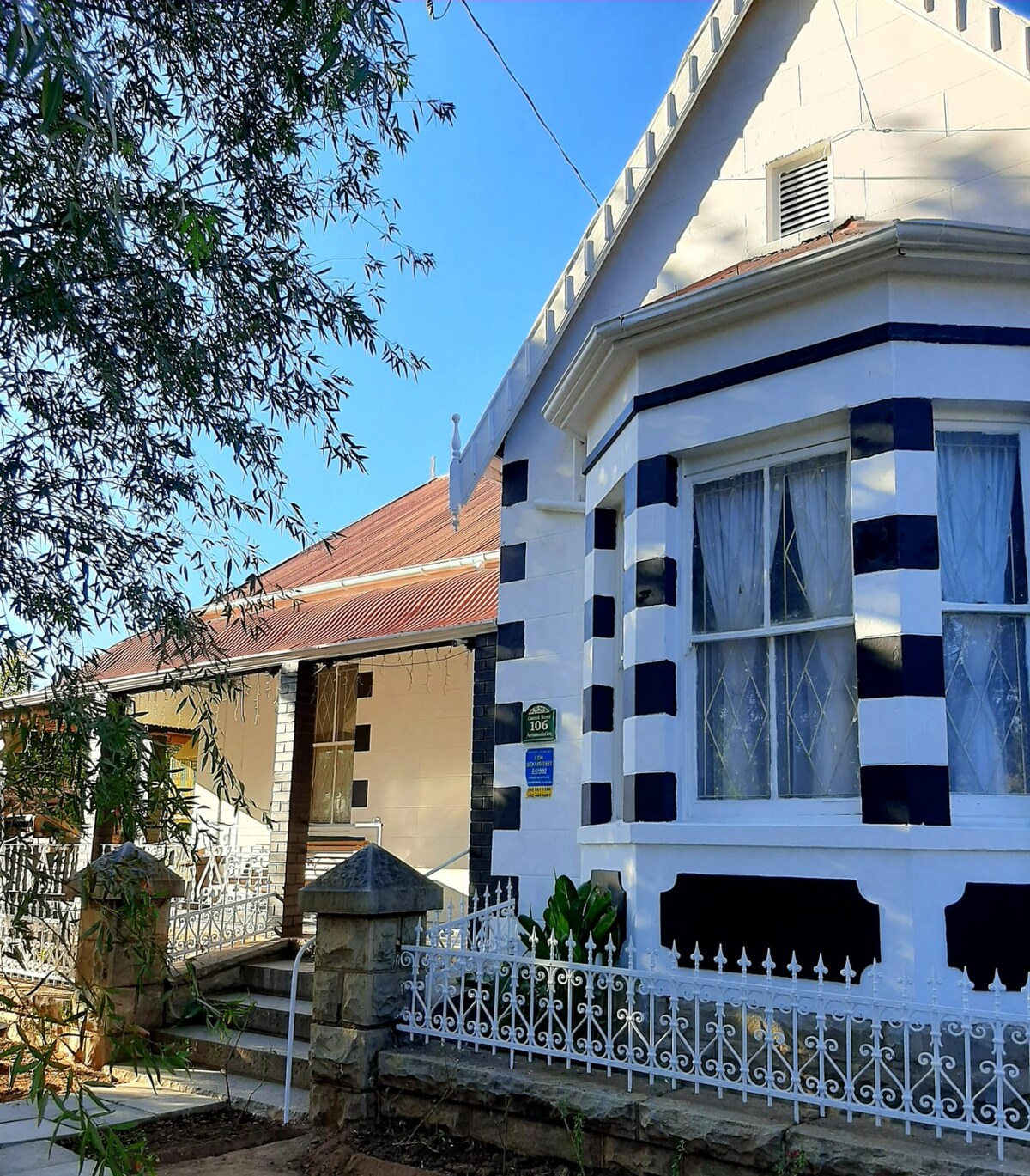 The exterior of a historic house features a unique combination of white and dark blue patterned walls. The entrance is framed by a decorative iron fence and lush green plants, with large windows adorned by sheer curtains allowing natural light to brighten the space.