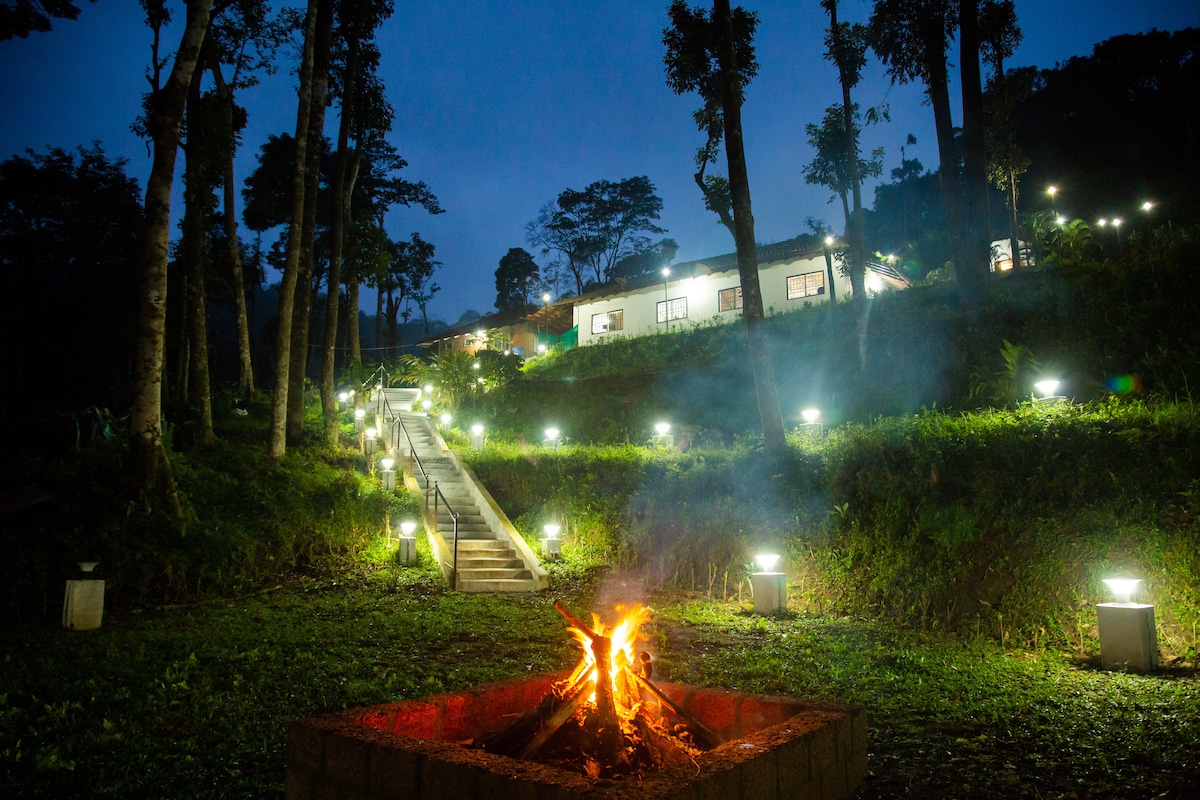 A warm fire glows in a stone pit, surrounded by lush greenery in a serene night setting. A well-lit pathway leads up steps to the cottages, framed by trees. Soft lights illuminate the area, enhancing the peaceful atmosphere of the hillside retreat.