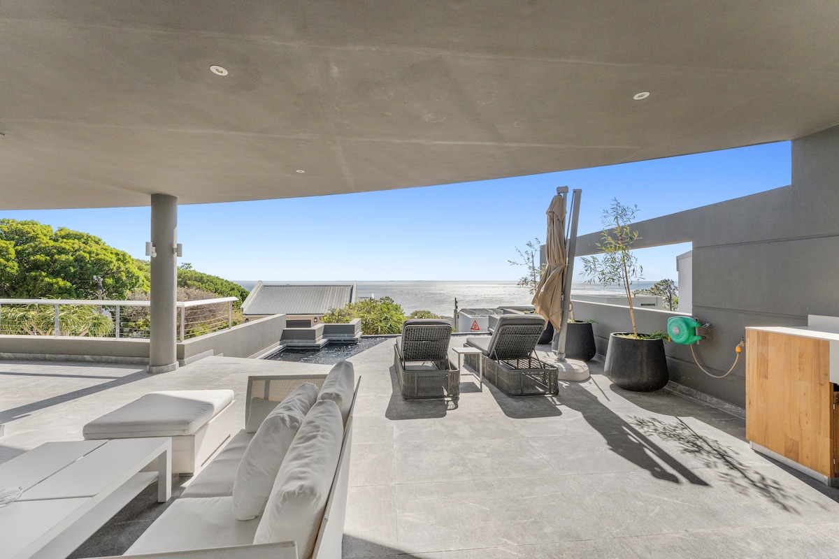 An outdoor terrace features a sun-drenched seating area with modern lounge furniture. A view of the ocean is visible beyond the space, while an infinity pool adds a touch of luxury. A gas braai is positioned nearby, complemented by greenery that enhances the ambiance.