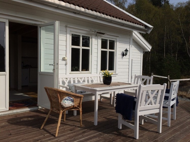 An outdoor dining area is presented with a wooden table surrounded by four white chairs. A single wicker chair adds seating. A potted plant is positioned on the table, and the entrance to the interior is visible through an open door.