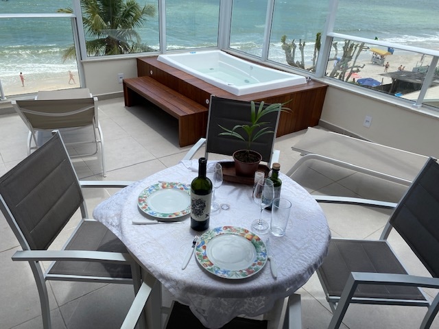 A dining table is set for two with plates and glasses, surrounded by comfortable chairs. In the background, a spa is visible alongside a view of the beach and ocean. Natural light enters through large windows, enhancing the serene atmosphere.