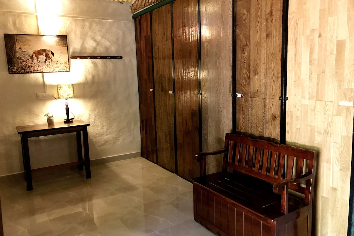The image presents a serene entryway featuring a wooden bench and a small table. Wooden cabinetry lines the wall, creating an organized appearance. Soft lighting from a lamp enhances the natural textures, while a framed image of wildlife adds a touch of nature-themed decor.