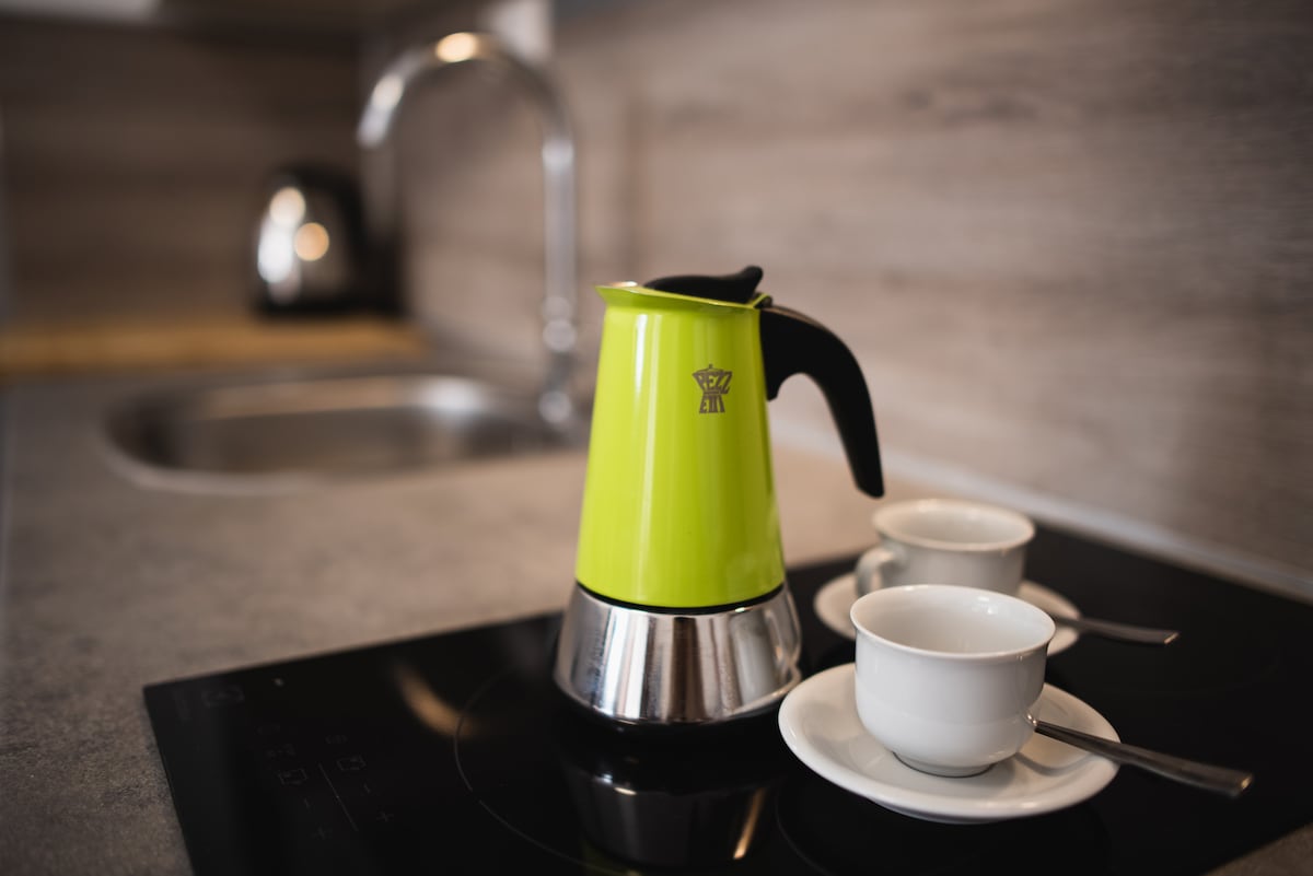 A bright green moka pot is positioned on a sleek stovetop, accompanied by two white espresso cups and small silver spoons. The setting features a modern kitchen design with subtle wood-textured walls and a polished countertop.