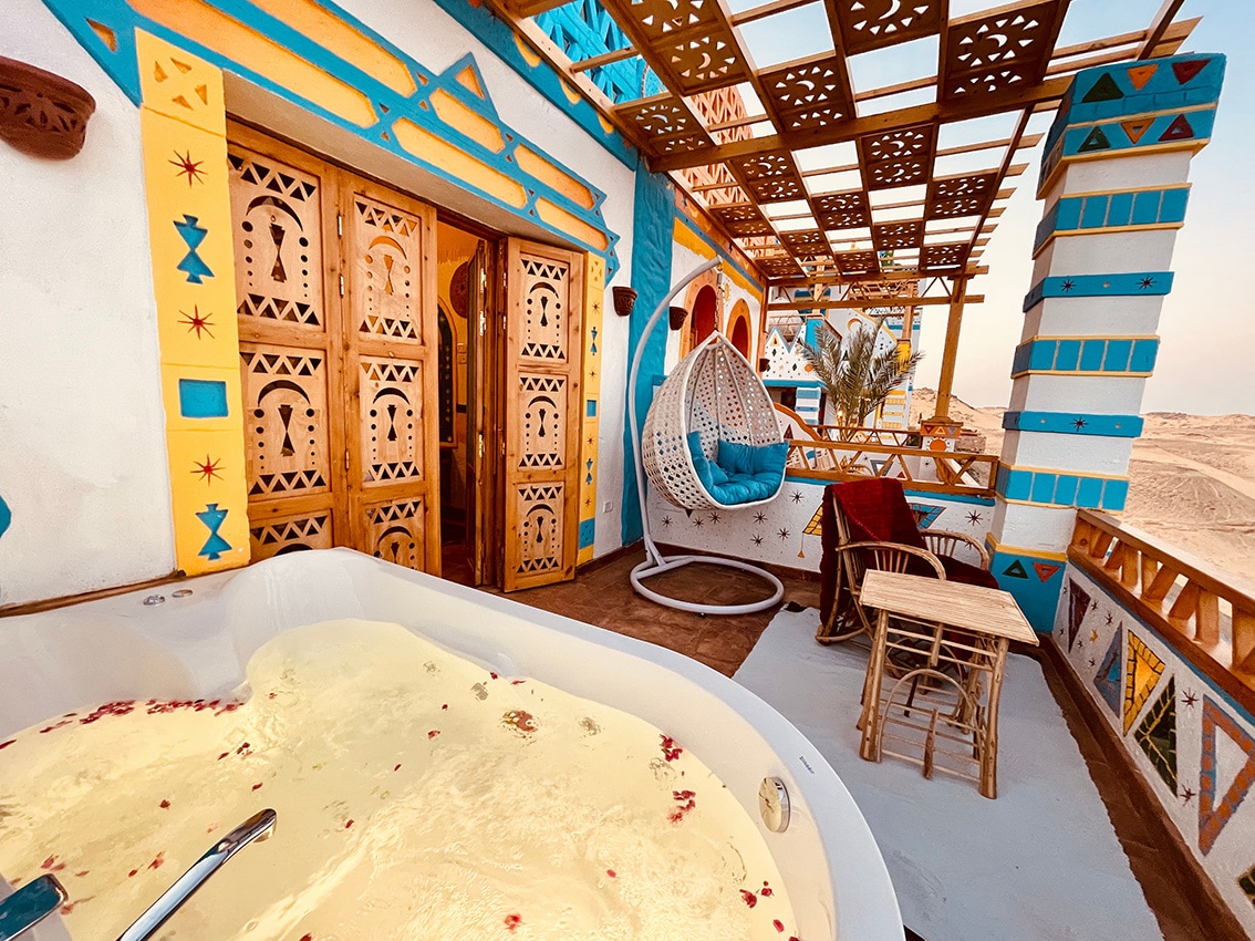 A private balcony features decorative blue and yellow walls, offering views of the surroundings. A hot tub filled with water and floating rose petals is visible, alongside a hanging chair and a small table with chairs. Ornate wooden doors add to the unique architectural detail.