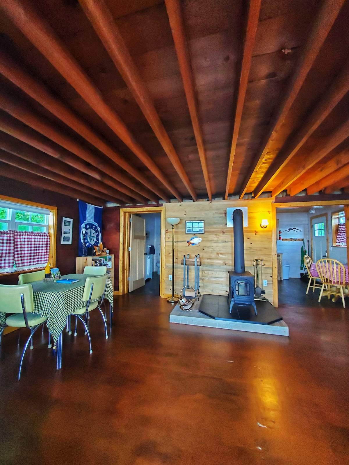 A warm living area features wooden ceiling beams and a wooden stove at the center. Bright yellow chairs surround a checkered table, while a woven rug lies on the polished floor. Light streams through windows adorned with checkered curtains, creating an inviting ambiance.