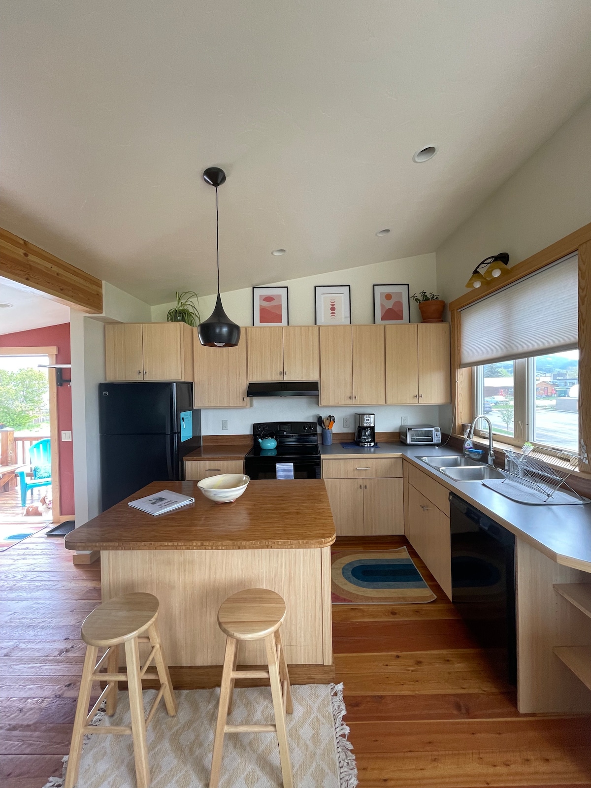 The kitchen features light wooden cabinetry and a central island with seating for two. Black appliances, including a stove and refrigerator, complement the space. Natural light filters through large windows, illuminating the counter areas, while decorative art hangs above the cabinets.