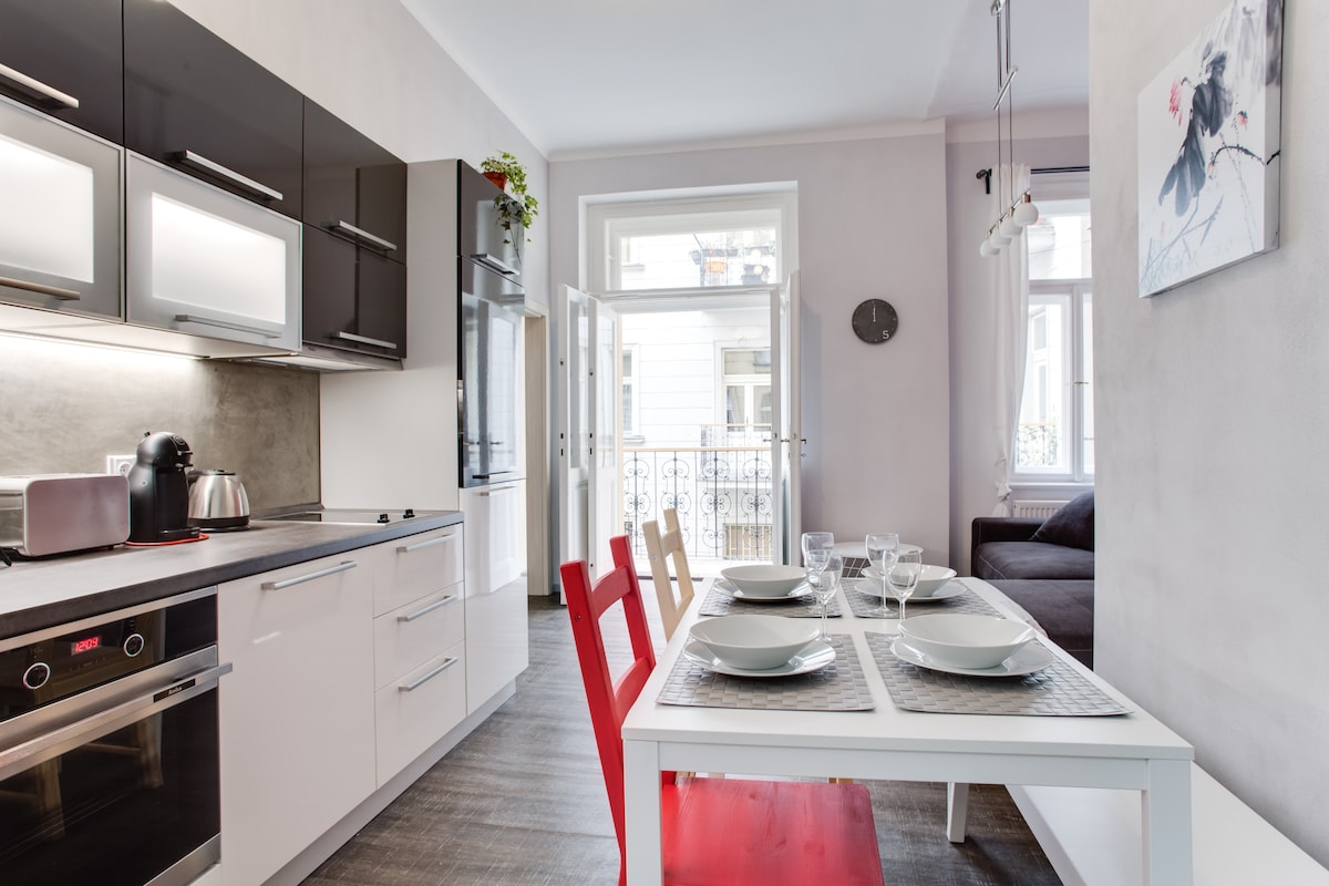 A modern kitchen features sleek cabinetry with integrated appliances, including a kettle and coffee machine. A dining table with red and white chairs accommodates six people and is set for a meal. Natural light filters in through the open balcony doors, illuminating the space.