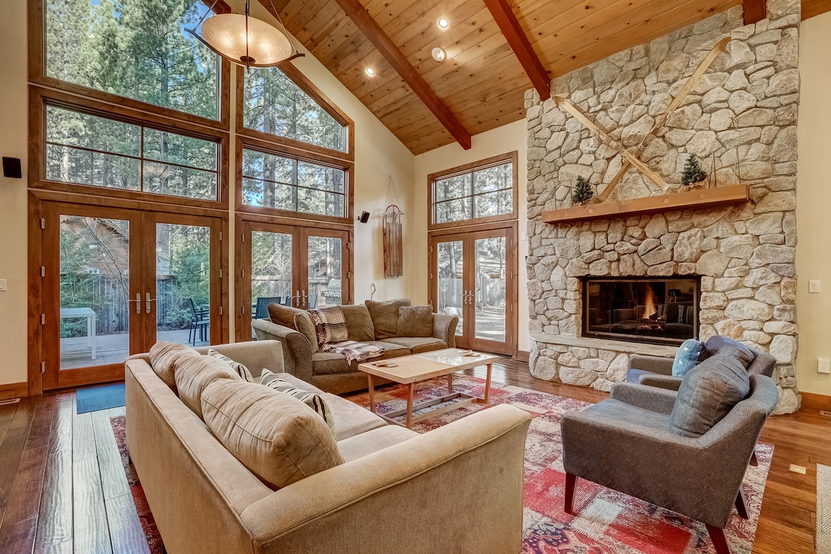A spacious living room features a high, vaulted ceiling with wooden beams. A stone fireplace serves as a central focal point, surrounded by large windows that fill the area with natural light. Comfortable seating arrangements include plush sofas and a coffee table, set against a richly patterned area rug.