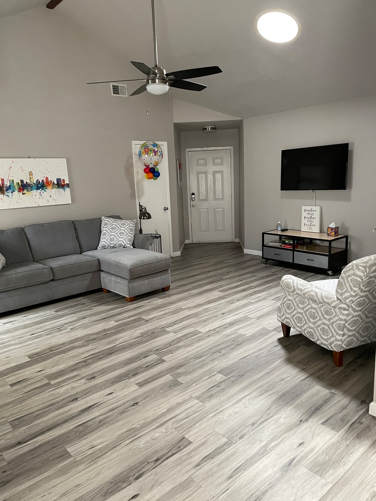 A living room features a comfortable gray sofa with decorative pillows and a matching armchair. A sleek television is mounted on the wall opposite the seating area. Light-colored flooring complements the neutral walls, while a colorful wall art piece adds a touch of personality.