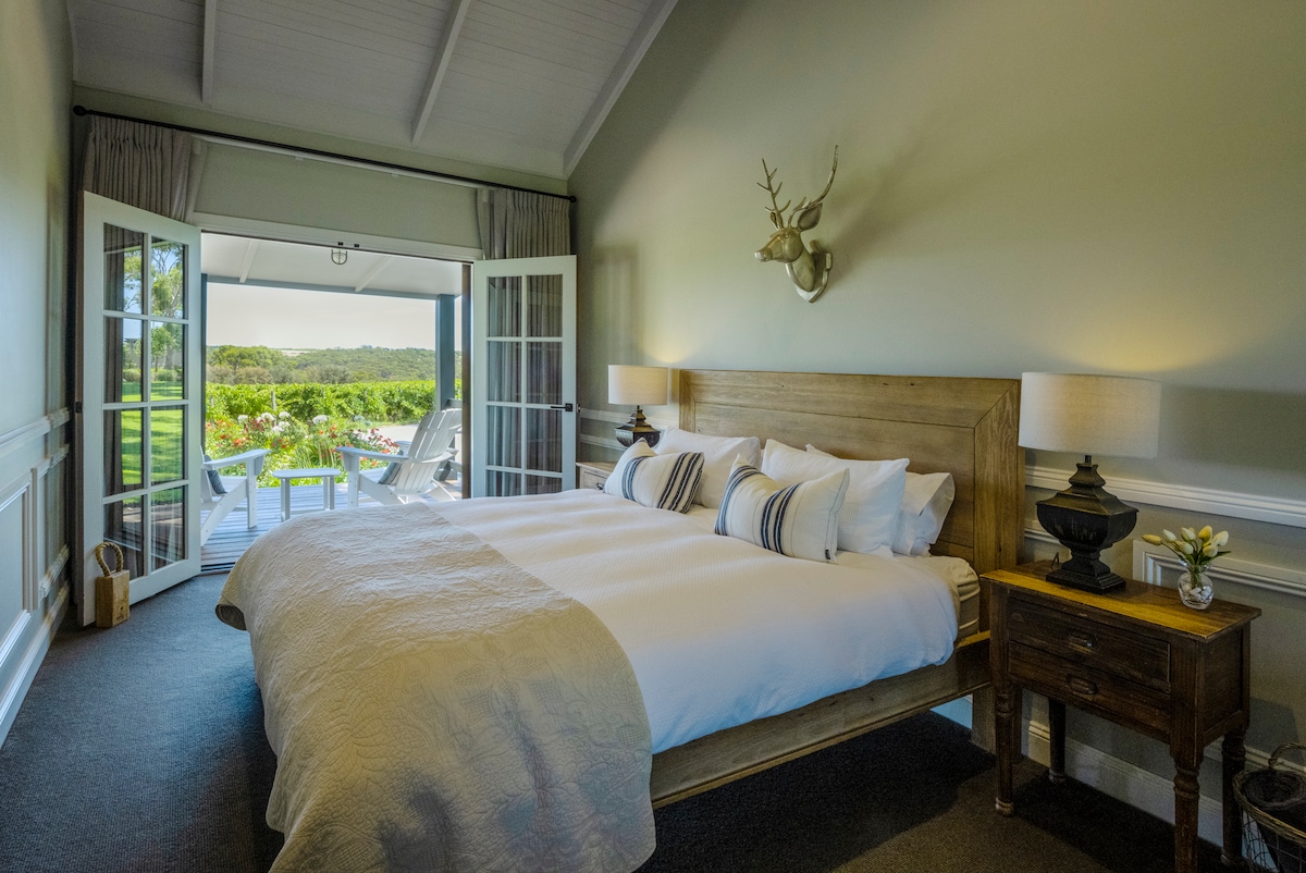 The spacious king bedroom features a large bed with luxury linen, complemented by bedside tables with lamps. Double doors open to a deck with vineyard views, allowing natural light to enhance the serene ambiance. The room is adorned with subtle decor, including a mounted antler piece.