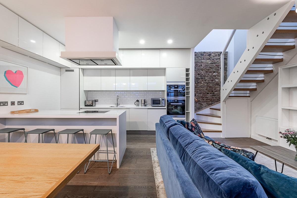 The open-plan kitchen and living area showcases sleek white cabinetry and a large island with seating for four. A wooden dining table is positioned to the side, while a comfortable blue sofa offers a cozy seating option. Natural light floods the space from the staircase leading to the upper level.
