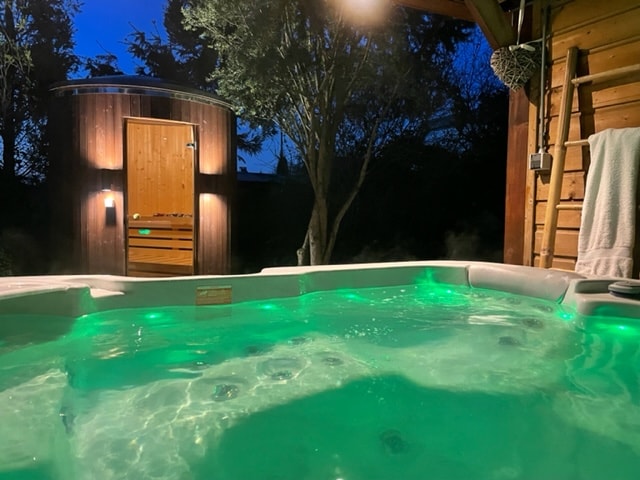 A secluded jacuzzi is illuminated by soft green lighting, offering a relaxing ambiance. The image also captures a wooden sauna structure in the background, surrounded by trees under a night sky. Steam rises gently from the water, enhancing the tranquil atmosphere.