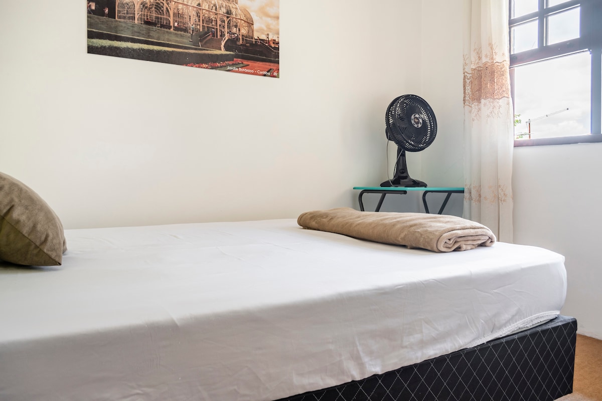 A clean and spacious sleeping area is featured, containing a neatly made bed with a light-colored fitted sheet and a rolled towel. A pedestal fan rests on a small table beside the bed, while natural light enters through a window adorned with sheer curtains.