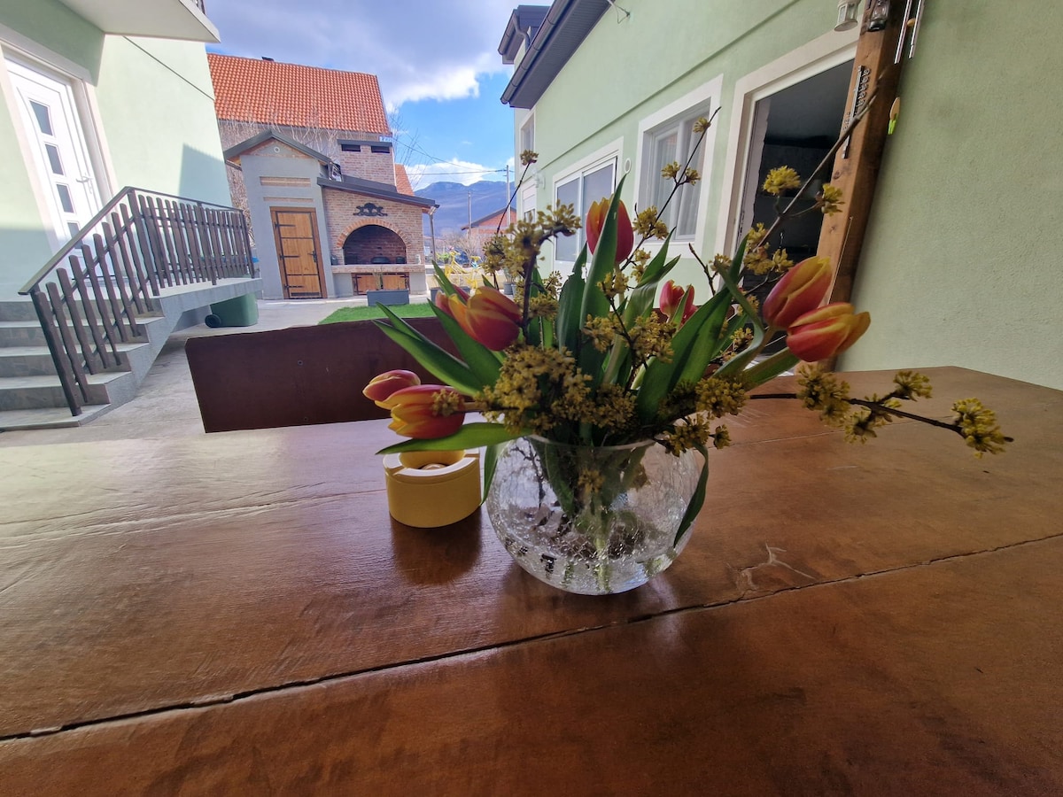 A clear glass vase holds a bouquet of tulips and yellow flowers on a wooden table. The background features a glimpse of a rustic building and outdoor space, with soft natural lighting enhancing the scene.