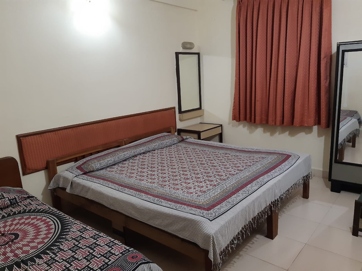 An air-conditioned bedroom features a spacious double bed and a single bed, both dressed with patterned bed linens. A mirror and a bedside table are visible, alongside a window covered by floor-length curtains that filter natural light.