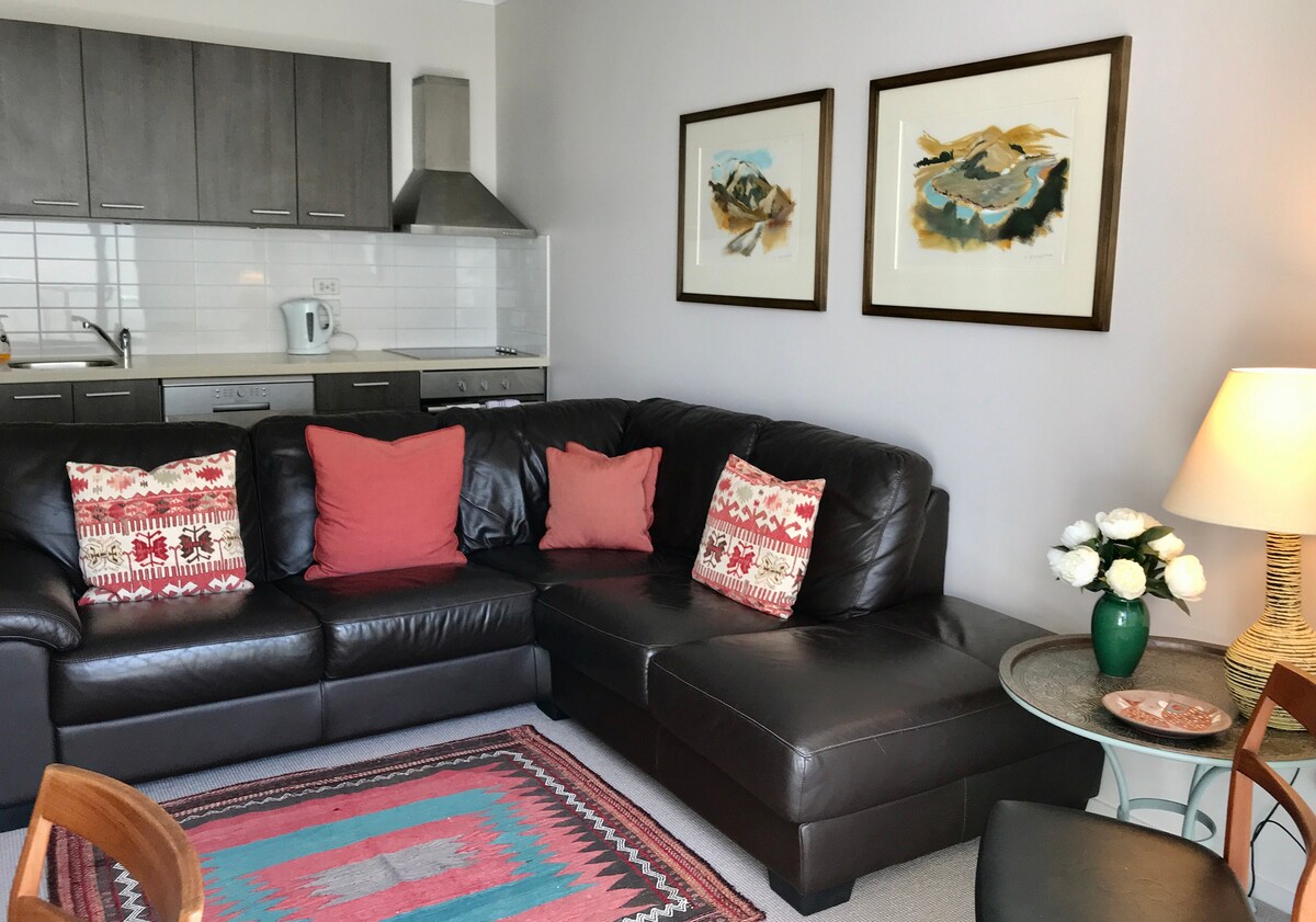 A spacious living area features an L-shaped leather sofa adorned with decorative cushions, complemented by a small side table holding a vase of flowers. Two framed artworks are displayed on the wall, and a kitchen area with modern appliances is partially visible in the background.