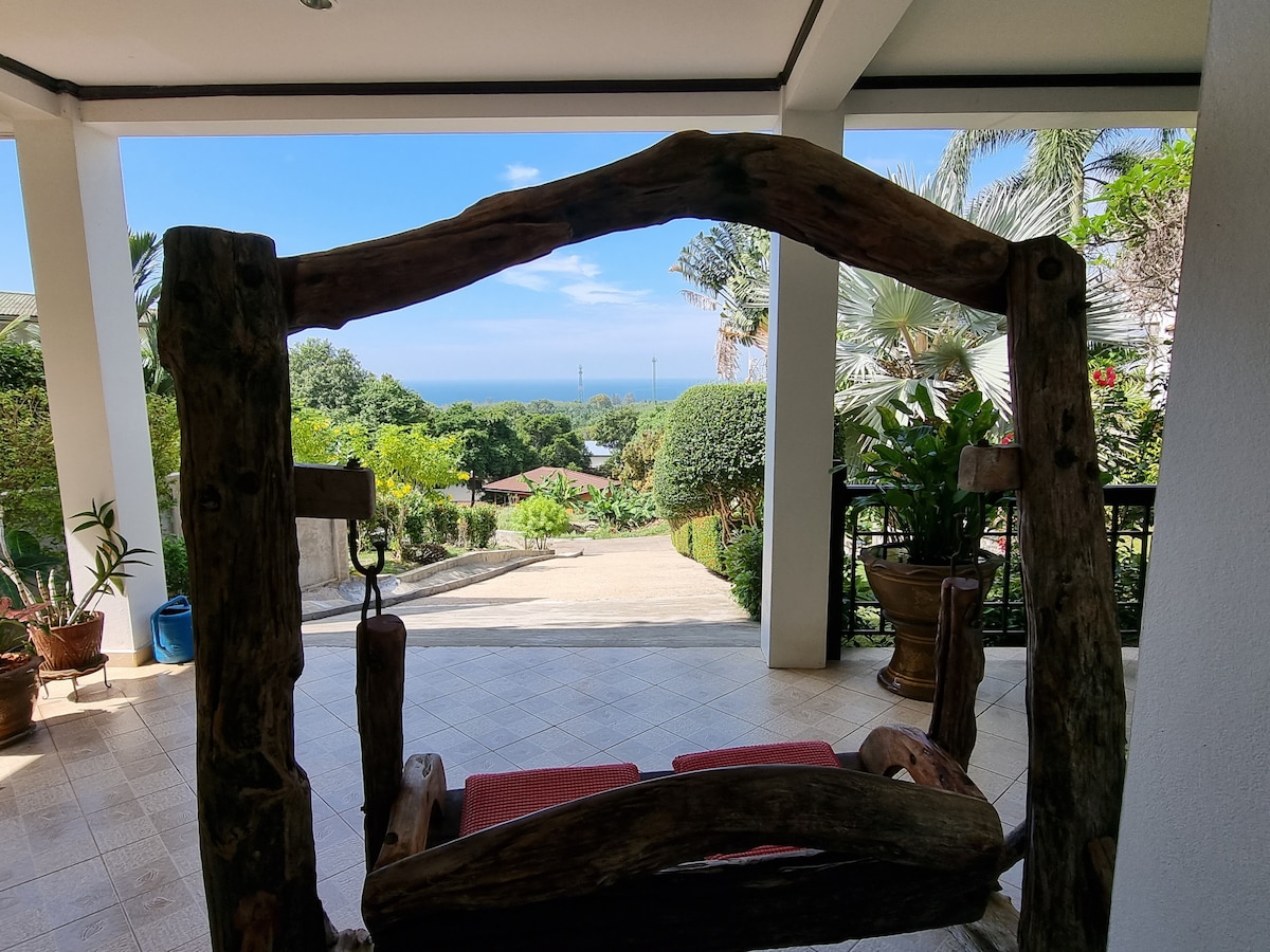 A wooden swing frame is positioned in the foreground, providing a view of the lush garden and the Andaman Sea in the background. The vibrant greenery surrounds the pathway that leads down from the terrace, framed by potted plants and trees.