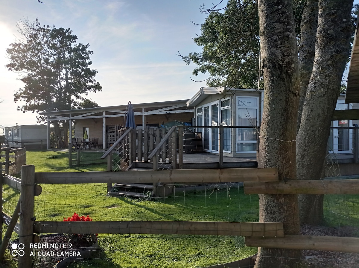 A mobile home is set on a private landscaped plot, featuring spacious outdoor decking and a staircase leading to the entrance. The area is surrounded by green grass and trees, with a flower bed adding a touch of color. Natural light highlights the structure's modern design.