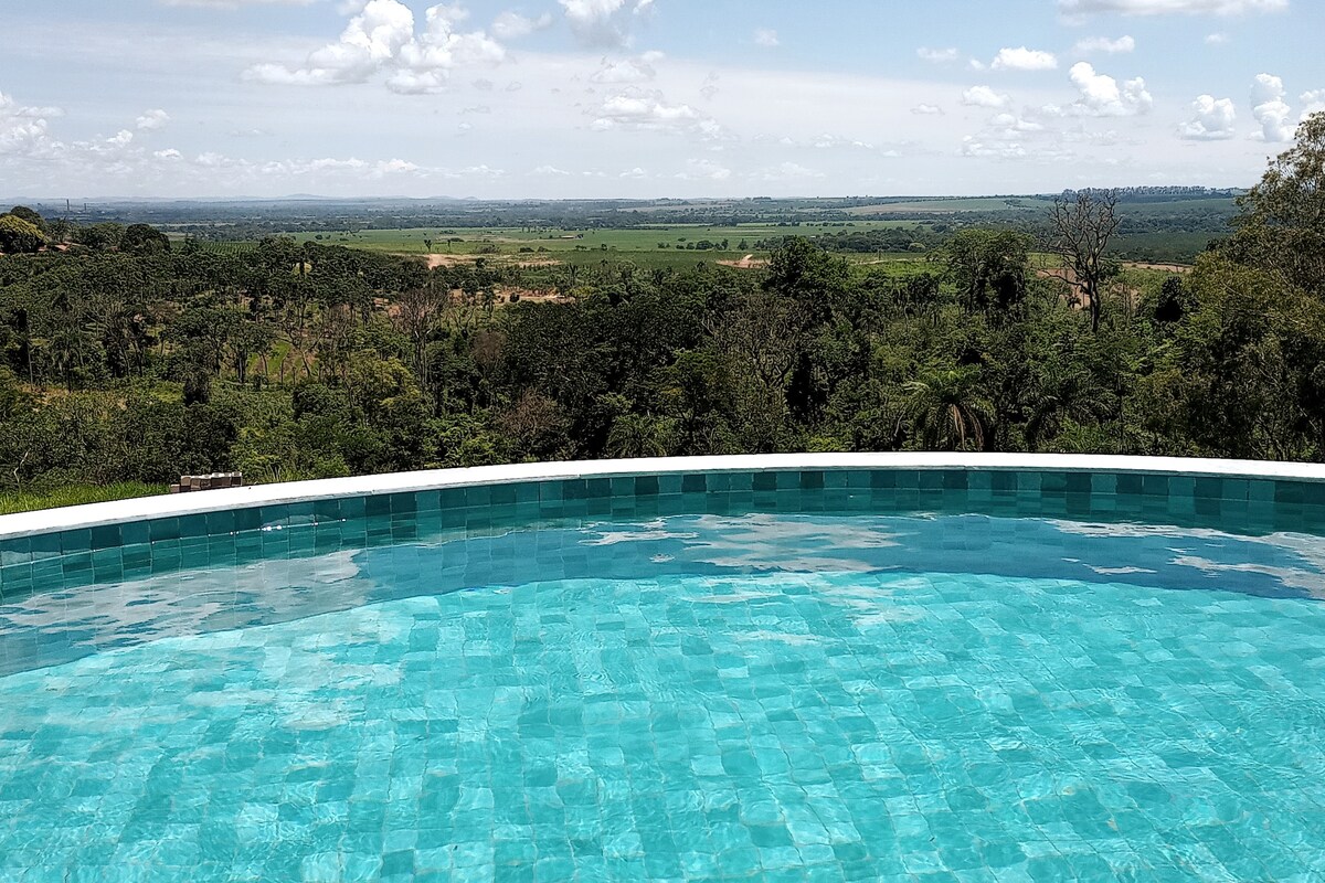 A clear swimming pool reflects the sky and surrounding landscape. The view encompasses lush greenery and distant hills, providing a serene backdrop. Sunlight creates a sparkling effect on the pool's surface, inviting relaxation and enjoyment of nature.