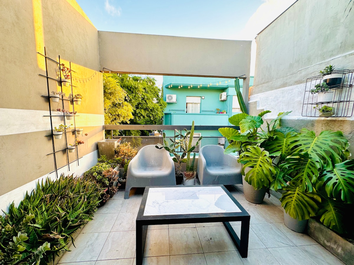 A terrace area is presented, featuring two grey lounge chairs positioned beside a black table. Various plants are displayed in wall-mounted and floor pots, creating a green oasis. Natural light brightens the space, offering a comfortable setting for relaxation and outdoor enjoyment.