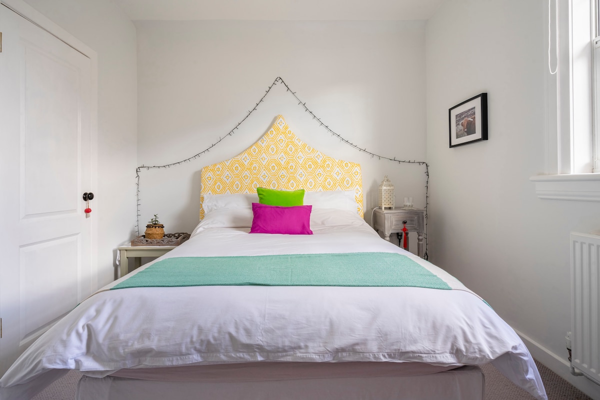 The bedroom features a spacious bed with a vibrant yellow headboard and colorful accent pillows. A soft light green throw covers the bed, and decorative fairy lights are draped along the wall. A small bedside table with a decorative item adds to the inviting ambiance.