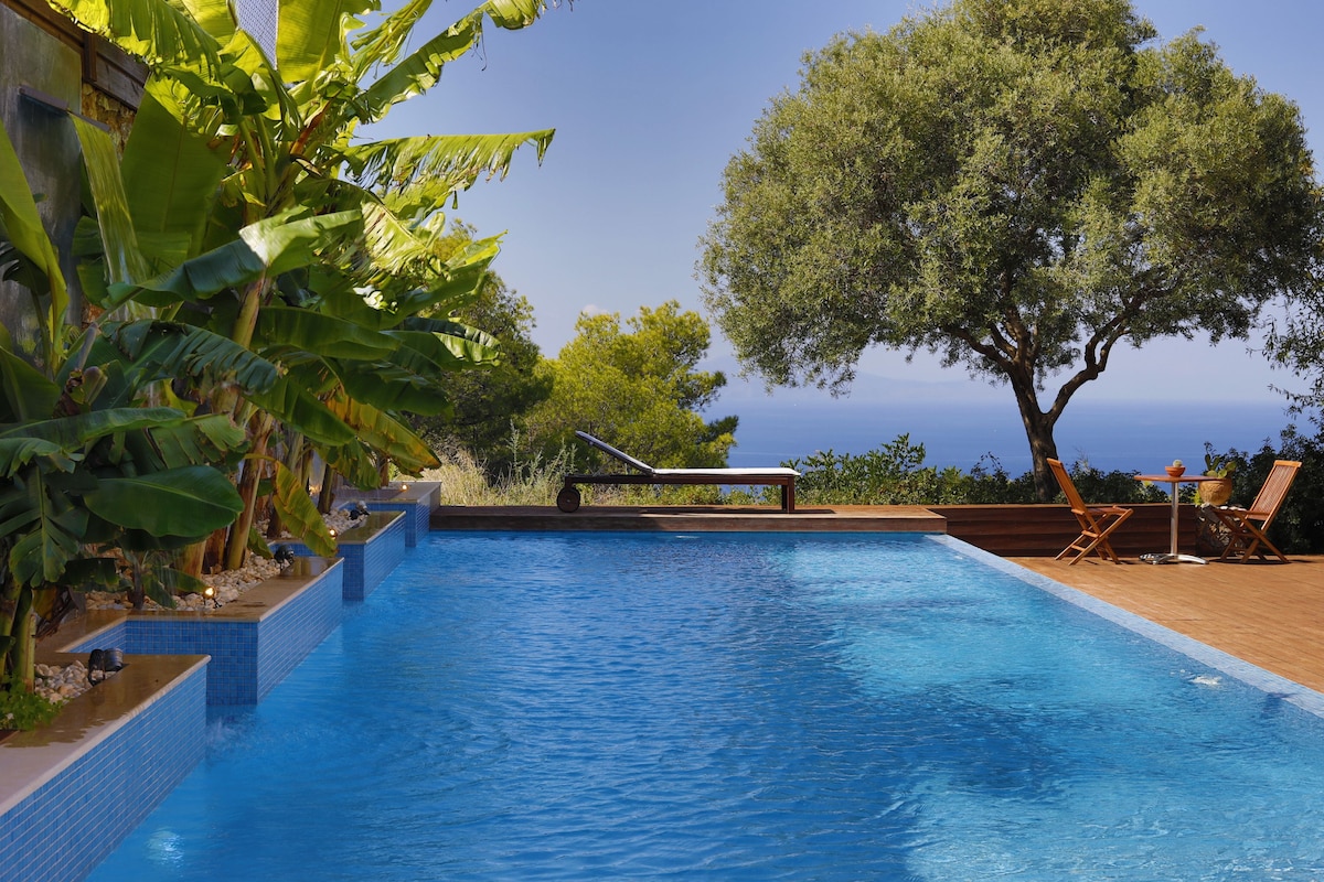 The outdoor area features a tranquil swimming pool surrounded by lush banana plants and an olive tree. A wooden deck provides additional lounging space, complemented by a small table and chairs inviting relaxation while enjoying views of the landscape and the sea.