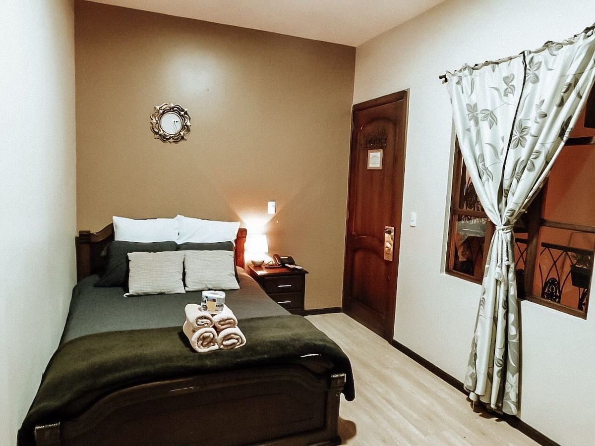 A cozy private room features a spacious bed adorned with soft pillows and a neatly folded set of towels. A bedside lamp provides illumination, while a window with elegant curtains allows soft light to enter. A wooden door leads to the hallway.