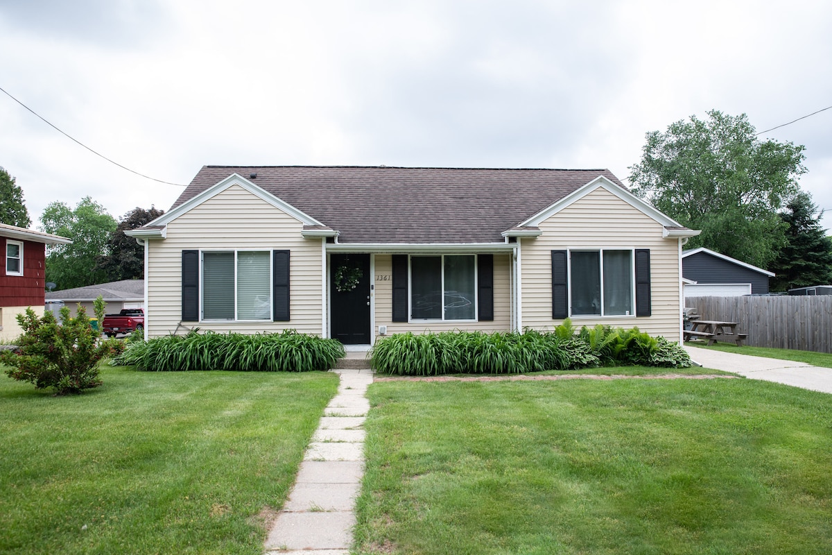 The front exterior of a single-story house is visible, featuring a tan facade with dark shutters and large windows. A well-maintained lawn surrounds the home, with green shrubs in front. A concrete pathway leads to the entrance, framed by an inviting green landscape.