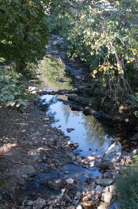 A rocky stream meanders through a natural setting. Lush green foliage frames the water, which reflects the trees and sky. The ground features scattered stones and pebbles, contributing to the serene rural atmosphere of the surrounding landscape.