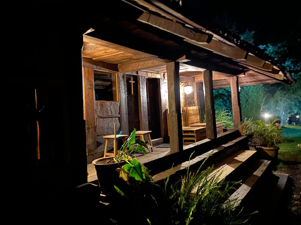 The rustic wooden cabin is presented at night, illuminated by warm lighting. A covered porch features two wooden chairs and a small table. Surrounding greenery adds to the natural ambiance, while the entrance is accessed via a few wooden steps.