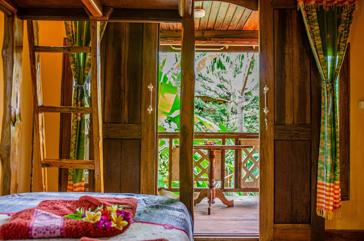 A spacious bedroom featuring natural wooden elements and an inviting atmosphere. Large double doors open to a generous balcony overlooking greenery. A cozy bed is adorned with a colorful blanket and decorative flowers. A ladder leads to additional upper spaces, enhancing accessibility.