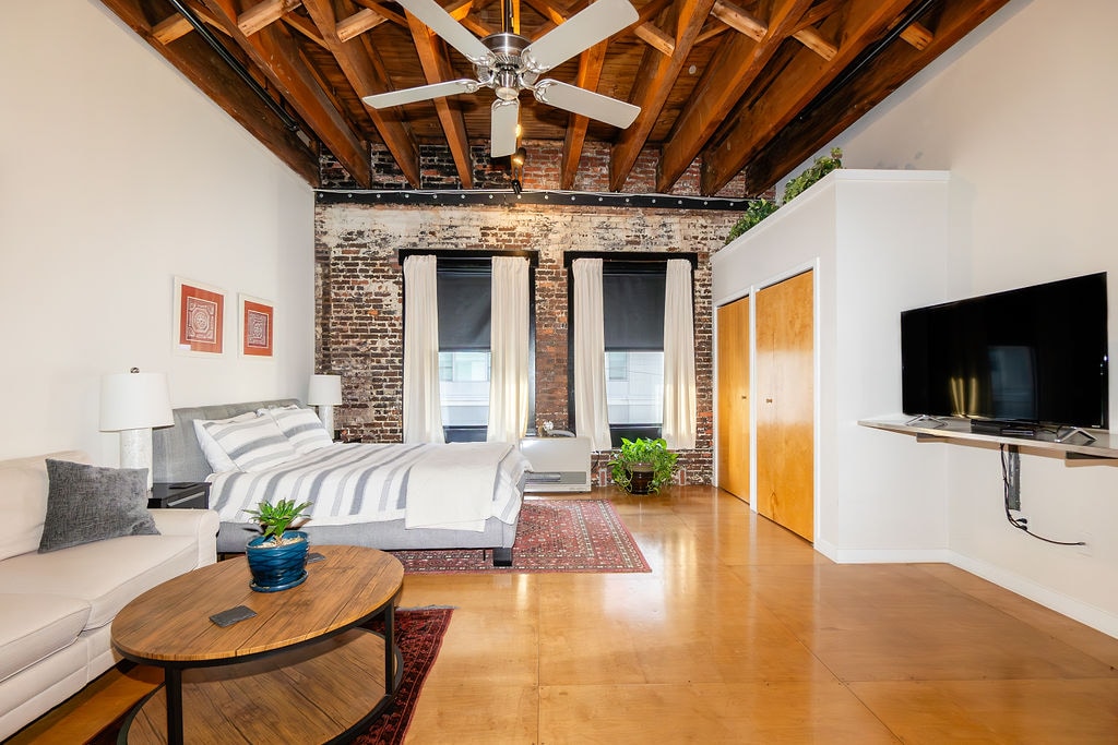 A spacious studio loft is illuminated by natural light, revealing exposed brick walls and wooden beams. A comfortable bed with striped bedding is positioned beside a modern television mounted on the wall. A cozy seating area includes a light-colored sofa, accompanied by a round coffee table and indoor plants.