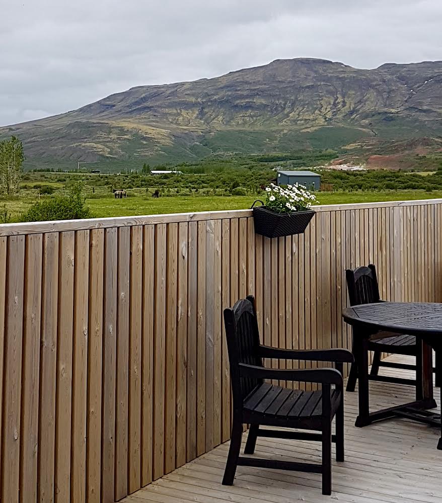 A wooden deck area is presented with black outdoor chairs and a round table. A planter with white flowers hangs from the wooden railing, while a green mountainous landscape is visible in the background, offering a serene and peaceful view.