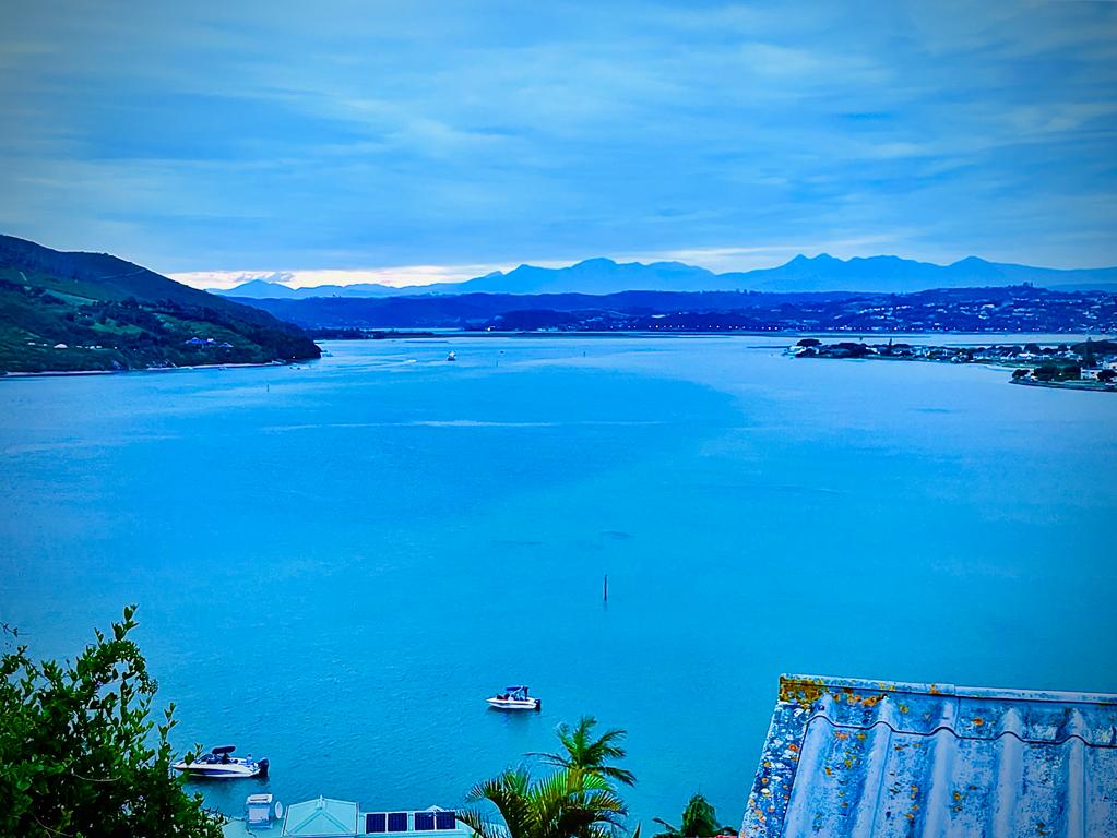 A wide view of a serene body of water reflects shades of blue, framed by distant hills and mountains. Boats can be seen floating, while a gentle shoreline enhances the scene. The sky appears overcast, adding a soft light to the tranquil landscape.