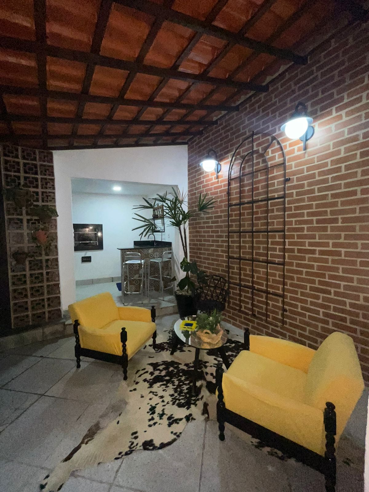 An outdoor seating area features two yellow chairs positioned around a glass table. A decorative wall with a plant adds greenery to the space. Soft lighting from two wall-mounted fixtures illuminates the area, creating a cozy environment sheltered by a textured ceiling.